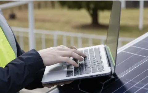 Praca nad projektem fotowoltaiki: ręce na klawiaturze laptopa, obok panel słoneczny.