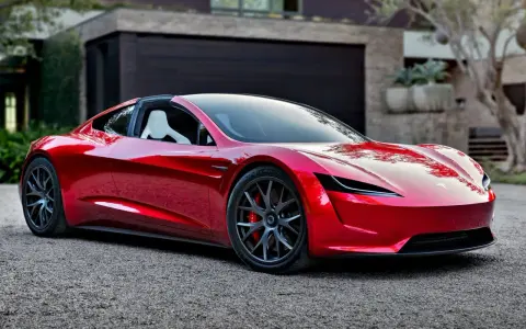 Błyszczący, czerwony Tesla Roadster 2 z białymi siedzeniami stoi przed nowoczesnym domem.