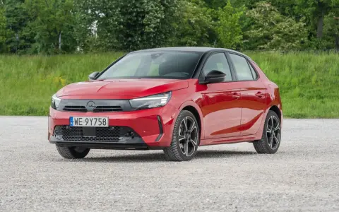 Czerwony Opel Corsa E na tle zieleni. Zbliżenie na przód auta, gdzie widoczne są światła LED i tablica rejestracyjna. Poznaj opel corsa e wymiary.