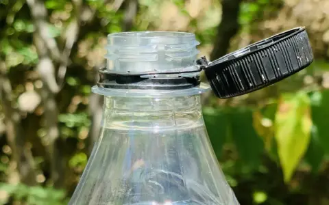 Nakrętki od butelek są przytwierdzone, by zapobiec ich zgubieniu i ułatwić ponowne zakręcenie. Widoczny fragment plastikowej butelki z otwartą, czarną nakrętką.