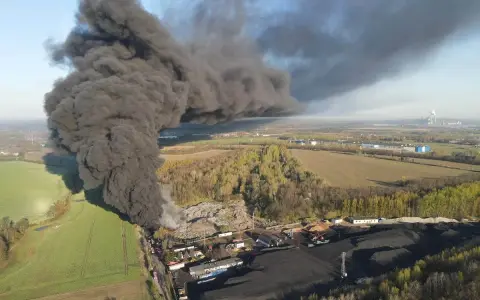 Ogromny słup czarnego dymu unosi się nad składowiskiem opon, gdzie trwa pożar. Widać wozy strażackie i budynki przemysłowe.