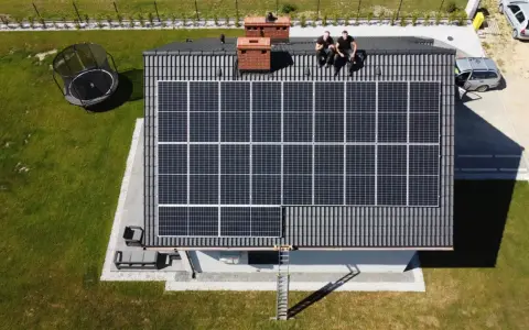 Dwaj mężczyźni siedzą na dachu domu z panelami słonecznymi, być może omawiając umowę rezerwową Tauron. Obok trampolina i samochody.