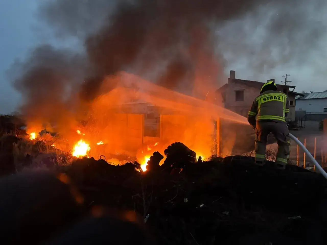 Strażak walczy z pożarem składowiska opon, z którego wydobywa się gęsty dym i płomienie.